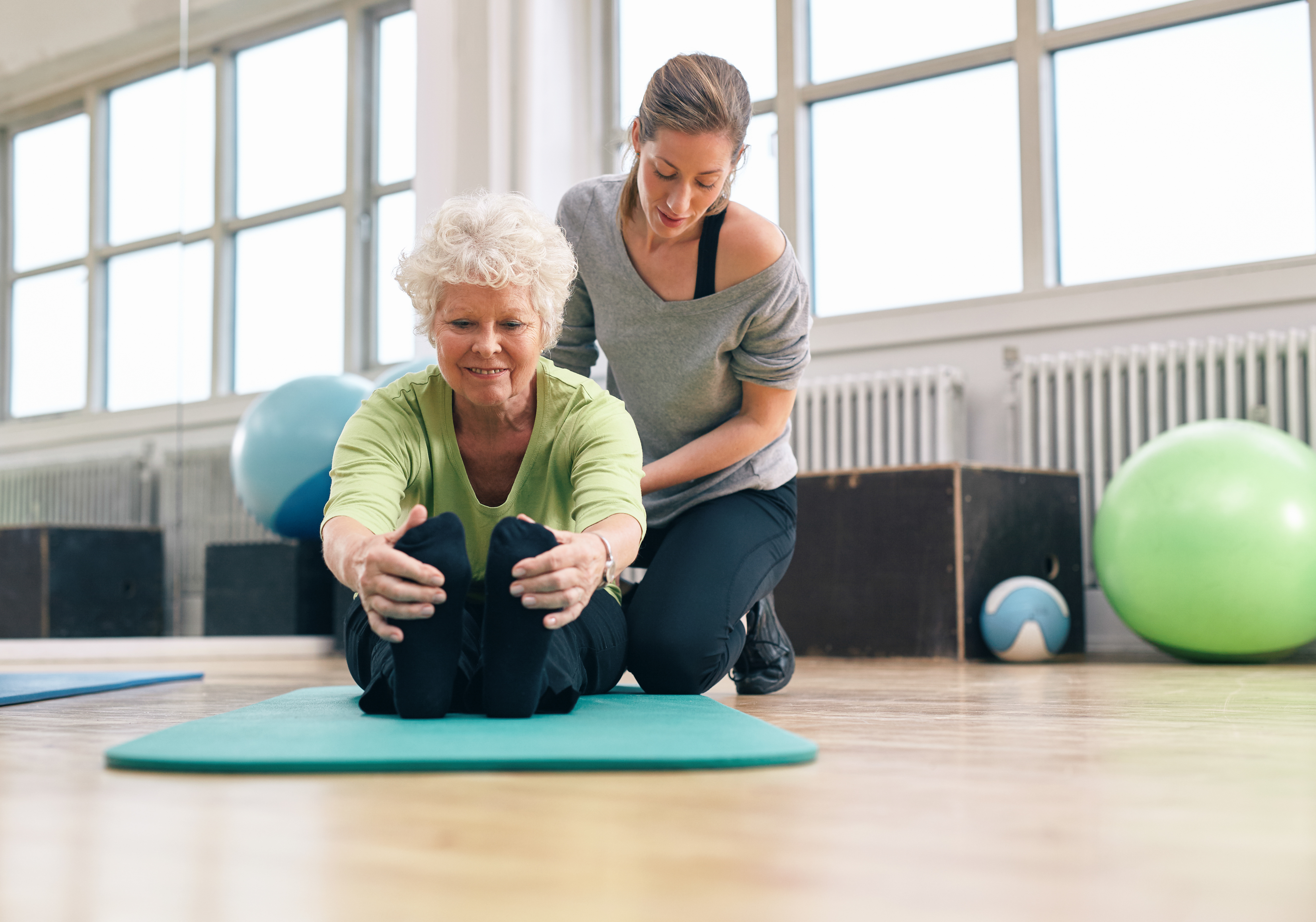 Elderly woman being helped by her instructor in the gym for exercising. Senior woman sitting on fitness mat bending forward and touching her toes with her personal trainer assisting.